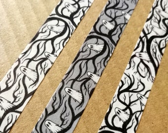 May include: Three rolls of black and white patterned washi tape with a spooky ghost and tree design. The tape is on a brown cardboard surface.
