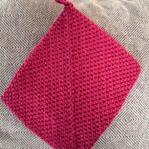 May include: A handmade, hot pink crochet potholder with a loop for hanging. The square-shaped potholder is made with a tight stitch pattern and a diagonal seam. The potholder is resting on a patterned, neutral-colored surface.