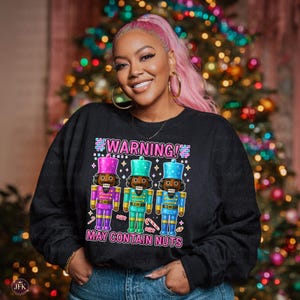 May include: Black sweatshirt with a graphic of three colorful nutcrackers. The text above the nutcrackers reads "WARNING!" and below reads "MAY CONTAIN NUTS". The nutcrackers are in purple, teal, and blue with candy canes.
