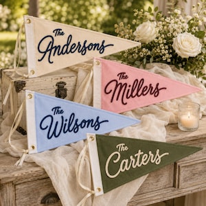 May include: Four triangular felt pennants with white canvas headers and brass grommets. The cream pennant reads The Andersons in navy, the pink reads The Millers in maroon, the blue reads The Wilsons in navy, and the olive green reads The Carters in cream.