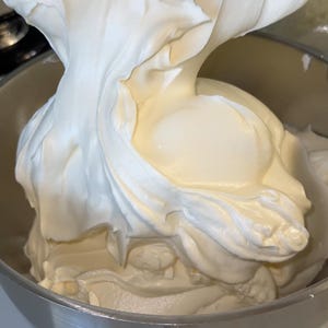 May include: Close-up of a stainless steel bowl filled with a large amount of white, whipped cream. The cream is being poured into the bowl, creating a swirl effect. The texture is smooth and fluffy.