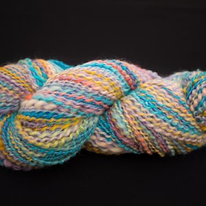 Peut inclure: Une pelote de laine multicolore avec des nuances de bleu, jaune, rose et violet. La laine est épaisse et texturée, idéale pour les projets de tricot ou de crochet. Le fond est noir uni.