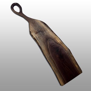 Puede incluir: Una tabla de servir de madera marrón oscuro con borde natural y asa con abertura circular. La tabla está hecha de una sola pieza de madera, mostrando la veta.