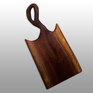 May include: A rectangular wooden cutting board with a handle. The board is made of dark brown wood with a natural grain pattern. The handle has a unique loop design. The board is angled, showing the wood's texture.