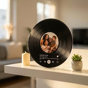 Könnte beinhalten: Eine schwarze Schallplatte mit einem Familienfoto in der Mitte. Die Platte zeigt den Songtitel "Forever Love" und den Künstlernamen "The Harmonizers". Ein weißes Regal hält die Platte, eine Kerze und eine kleine Topf-Sukkulente.