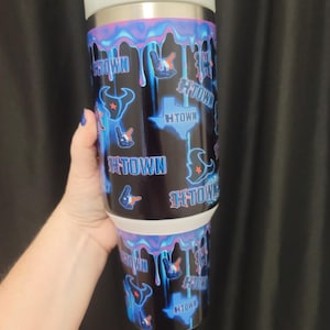 Puede incluir: Un vaso de acero inoxidable con tapa blanca y base negra. El vaso está decorado con un diseño de goteo azul y morado, junto con el texto "H-TOWN", un contorno de Texas y gráficos temáticos de béisbol.