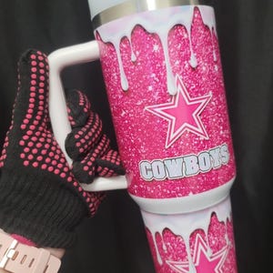 Könnte beinhalten: Ein pinkfarbener Glitzerbecher mit weißem Griff und Deckel. Der Becher hat ein weißes Tropfendesign und ein pinkfarbenes Sternlogo mit dem Wort "COWBOYS". Der Becher wird von einem schwarz-rosa Handschuh gehalten.