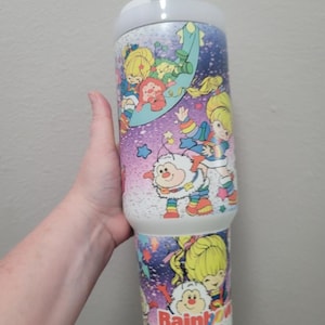 Può includere: Un bicchiere Rainbow Brite bianco e blu con personaggi dei cartoni animati e la scritta "Rainbow Brite". Il bicchiere ha un coperchio bianco e una forma cilindrica. Il design include stelle, arcobaleni e vari personaggi in uno stile colorato e stravagante.