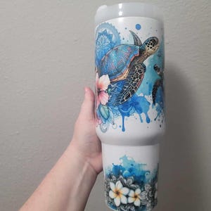May include: White tumbler with a colorful sea turtle design. The tumbler features a blue, pink, and green turtle illustration with floral accents and blue watercolor splashes. The bottom of the tumbler has a floral design.