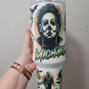 Könnte beinhalten: Weißer Becher mit einem Design von Michael Myers aus der Halloween-Filmreihe. Das Design beinhaltet den Namen "MICHAEL" in einem Graffiti-Schriftstil, Fledermäuse und andere Horror-Illustrationen. Der Becher hat einen weißen Deckel.
