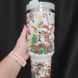 Könnte beinhalten: Ein weißer Edelstahlbecher mit einer bunten, weihnachtlichen Grafik der Figur Gizmo aus Gremlins mit Weihnachtsmannmütze, umgeben von Weihnachtsbäumen, verpackten Geschenken, Zuckerstangen und Pfefferminzbonbons, inklusive eines weißen Deckels.