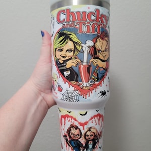 Puede incluir: Un vaso térmico blanco y plateado con el texto "Chucky and Tiff" e imágenes de dibujos animados de Chucky y Tiffany. El diseño incluye corazones, arañas, murciélagos y otros elementos temáticos de terror.