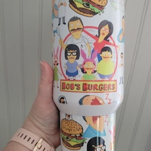May include: A white insulated travel mug with a colourful design featuring characters and burgers from the animated series "Bob's Burgers." The mug has a white lid. The text "Bob's Burgers" is visible on the mug.
