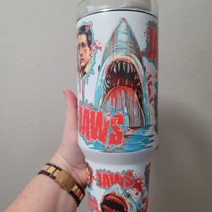 Puede incluir: Vaso blanco con un gráfico de un tiburón con la boca abierta, sangre y la palabra "JAWS" en rojo. El diseño incluye un retrato de un hombre y las palabras "JAWS" y "Jaws Gonna Get Ya".