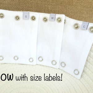 May include: Three white fabric pieces with metal eyelets and size labels marked S, M, and L. The text "NOW with size labels!" is at the bottom of the image. These are likely for clothing or accessories.