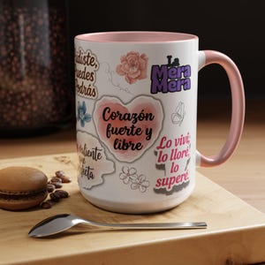 Peut inclure: Mug en céramique blanche avec intérieur et anse roses. Le mug est décoré de divers autocollants avec des motifs floraux et du texte espagnol, dont "Corazón fuerte y libre."