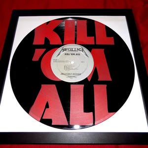 May include: Framed vinyl record with a black background and large red text that reads "KILL 'EM ALL". The record label in the center reads "METALLICA KILL 'EM ALL". The frame is black with a white mat.