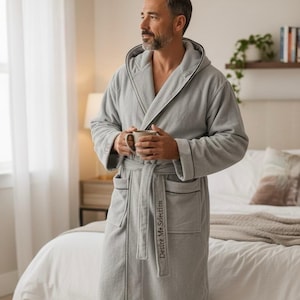 May include: A light grey hooded robe with black trim and a matching belt. The robe has pockets and the text "Desire Me Selection" on the belt. The person in the image is holding a mug.