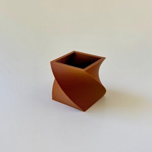 May include: A brown, square vase with a twisted design. The vase has a textured surface and a hollow interior. The overall shape is a combination of a square and a spiral, creating a unique and modern aesthetic.