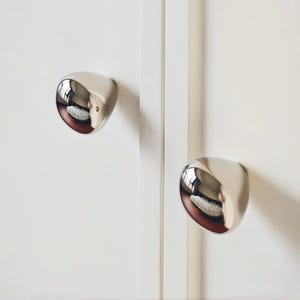 Peut inclure: Deux boutons de placard chromés et polis, à surface réfléchissante, montés sur une porte de placard blanche. Les boutons ont une forme arrondie en goutte d'eau. Le fond est une surface blanche unie.