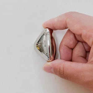 May include: A small, shiny, silver-colored object with a rounded, triangular shape. The object has a reflective surface and appears to be made of metal. The item is held between fingers against a white background.