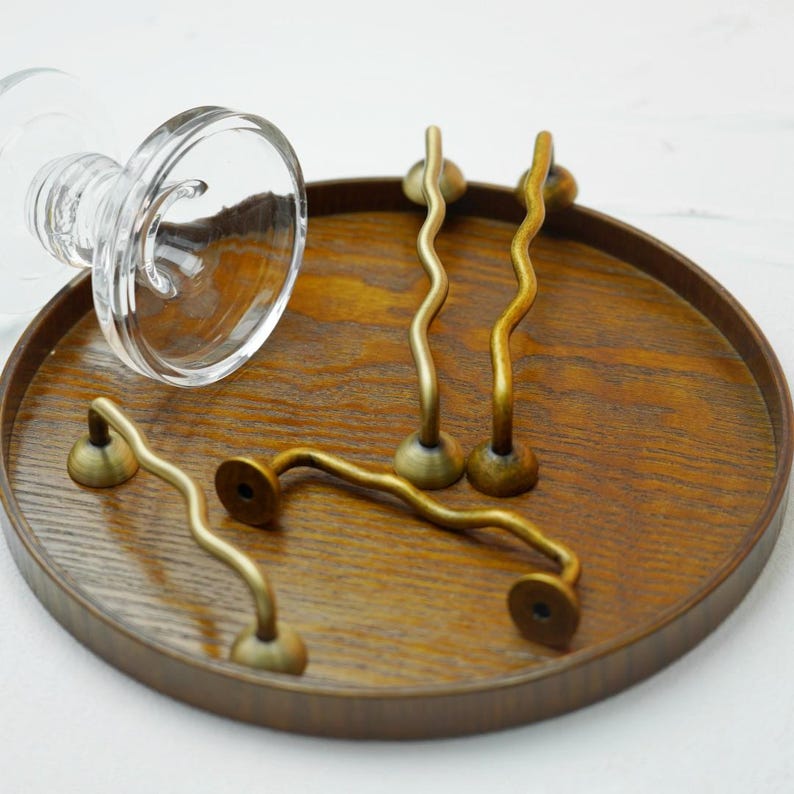 K&ouml;nnte beinhalten: Eine Sammlung von wellenf&ouml;rmigen Schrankgriffen in Messingoptik und ein klarer Glasgegenstand liegen auf einem runden Holztablett. Die Griffe haben ein einzigartiges, geschwungenes Design, und das Tablett hat einen dunkelbraunen Rand. Der Glasgegenstand hat einen ausgestellten Fu&szlig;.