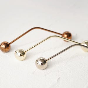 Pu&ograve; includere: Tre spille da bavero in metallo con teste sferiche. Una &egrave; color rame, una &egrave; dorata e una &egrave; argentata. Le spille hanno un design curvo e sono disposte diagonalmente su una superficie bianca.