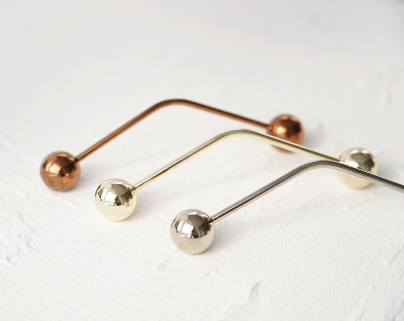 Pu&ograve; includere: Tre spille da bavero in metallo con teste sferiche. Una &egrave; color rame, una &egrave; dorata e una &egrave; argentata. Le spille hanno un design curvo e sono disposte diagonalmente su una superficie bianca.