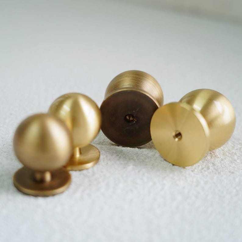 Op de afbeelding: Een verzameling van vijf messing kastknoppen. Elke knop heeft een afgeronde bol bovenop een platte, cirkelvormige basis. De knoppen hebben een warme, gouden tint, met kleine kleurvariaties. De hardware is ontworpen voor meubels en woondecoratie.