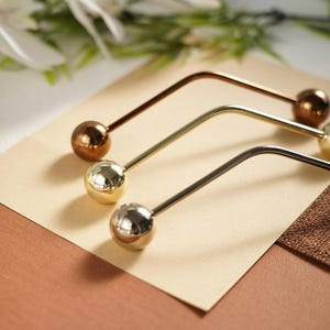 Pu&ograve; includere: Tre forcine per capelli in metallo bronzo, oro e argento. Ogni forcina ha un design curvo e un'estremit&agrave; sferica. Le forcine sono disposte su una superficie beige chiaro, con uno sfondo marrone e verde.