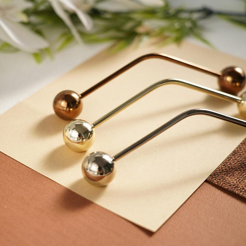 Pu&ograve; includere: Tre forcine per capelli in metallo bronzo, oro e argento. Ogni forcina ha un design curvo e un'estremit&agrave; sferica. Le forcine sono disposte su una superficie beige chiaro, con uno sfondo marrone e verde.