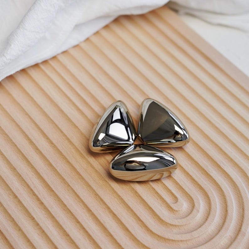 May include: Three shiny, metallic, triangular objects are arranged on a light brown, ridged wooden surface. The objects reflect light, creating a bright, polished appearance. A white cloth is visible in the background.