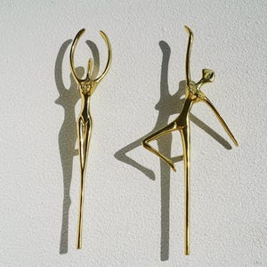 Puede incluir: Dos esculturas de metal dorado de bailarines estilizados. Una figura tiene los brazos levantados, la otra está en pose de ballet con una pierna extendida. Las esculturas están sobre un fondo blanco, proyectando largas sombras.