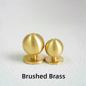 Op de afbeelding: Twee knoppen van geborsteld messing met een bolvormige bovenkant en een cirkelvormige basis. De knoppen zijn in verschillende maten en staan tegen een witte achtergrond. De tekst "Brushed Brass" staat eronder.