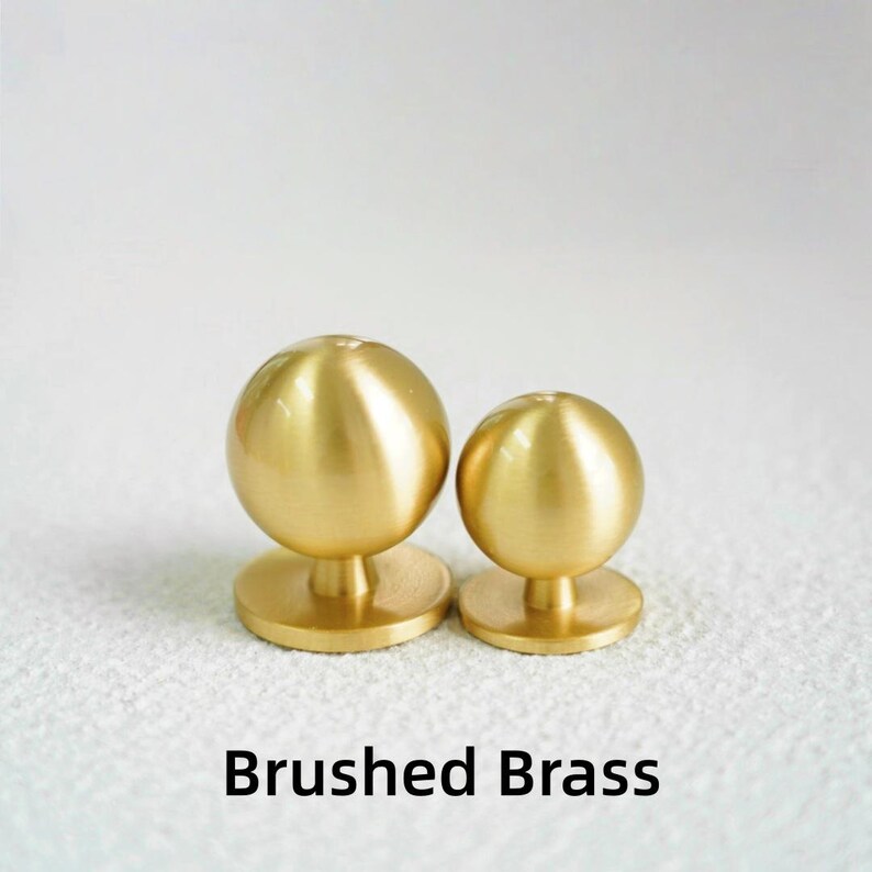 Op de afbeelding: Twee knoppen van geborsteld messing met een bolvormige bovenkant en een cirkelvormige basis. De knoppen zijn in verschillende maten en staan tegen een witte achtergrond. De tekst "Brushed Brass" staat eronder.