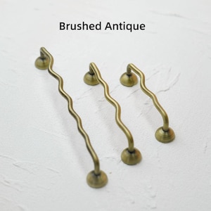 K&ouml;nnte beinhalten: Drei antike Messing-Schrankgriffe mit geb&uuml;rsteter Oberfl&auml;che und wellenf&ouml;rmigem Design. Die Griffe haben einen runden Sockel und einen gebogenen Griff, mit dem Text "Brushed Antique" oben in der Mitte.
