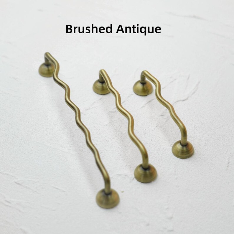 K&ouml;nnte beinhalten: Drei antike Messing-Schrankgriffe mit geb&uuml;rsteter Oberfl&auml;che und wellenf&ouml;rmigem Design. Die Griffe haben einen runden Sockel und einen gebogenen Griff, mit dem Text "Brushed Antique" oben in der Mitte.