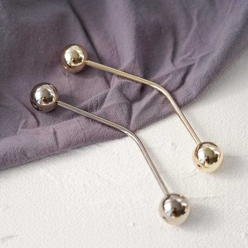 Pu&ograve; includere: Due spille a barra in metallo con estremit&agrave; sferiche. Una spilla &egrave; dorata, l'altra argentata. Le spille sono curve e poggiano su una superficie di tessuto grigio. Le spille vengono probabilmente utilizzate per fissare indumenti o accessori.