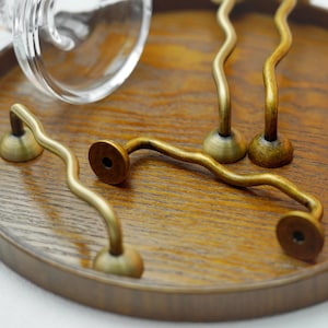 K&ouml;nnte beinhalten: Eine Sammlung antiker Messing-Schrankgriffe mit wellenf&ouml;rmigem Design. Die Griffe sind auf einem runden Holztablett angeordnet, das ihre einzigartige Form und das gealterte Finish zeigt. Die Hardware ist ideal, um M&ouml;beln einen Vintage-Touch zu verleihen.