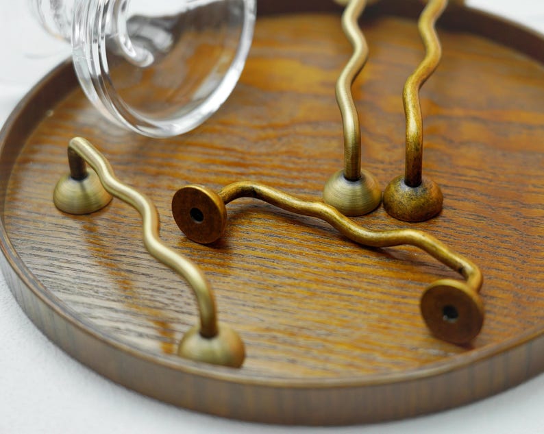 K&ouml;nnte beinhalten: Eine Sammlung antiker Messing-Schrankgriffe mit wellenf&ouml;rmigem Design. Die Griffe sind auf einem runden Holztablett angeordnet, das ihre einzigartige Form und das gealterte Finish zeigt. Die Hardware ist ideal, um M&ouml;beln einen Vintage-Touch zu verleihen.