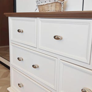 May include: A white dresser with multiple drawers and gold-toned knobs. The dresser has a dark brown top and is topped with a wicker basket with a polka-dot lining. The dresser has a classic, elegant design.