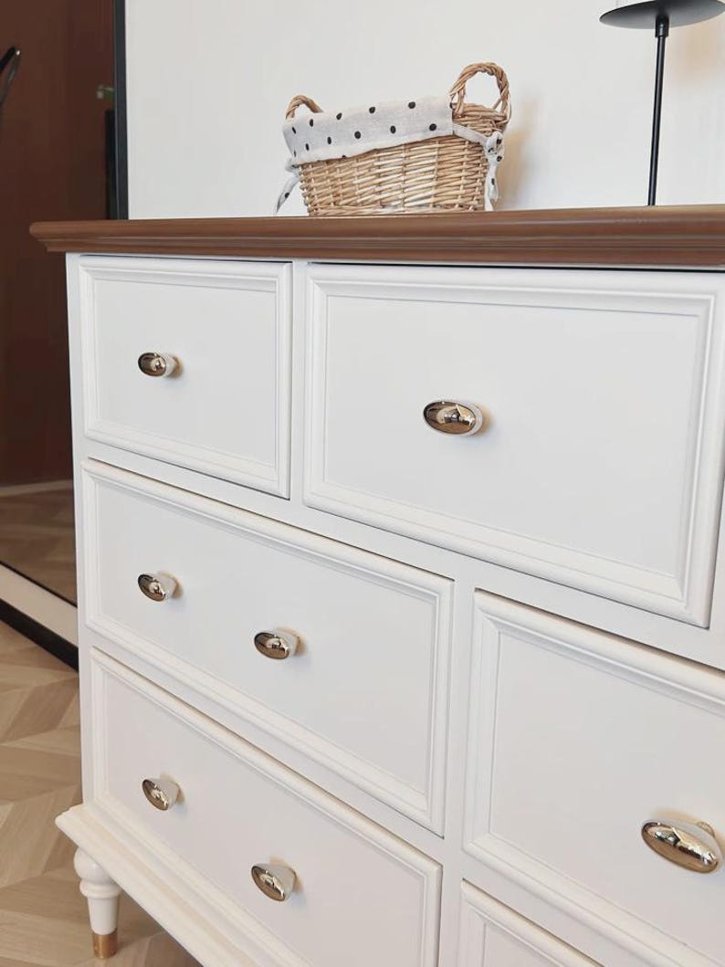 May include: A white dresser with multiple drawers and gold-toned knobs. The dresser has a dark brown top and is topped with a wicker basket with a polka-dot lining. The dresser has a classic, elegant design.