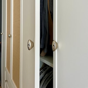 May include: A white wardrobe with gold-colored, cone-shaped knobs. The wardrobe doors are partially open, revealing clothing inside. Some doors have a woven, light brown panel design.