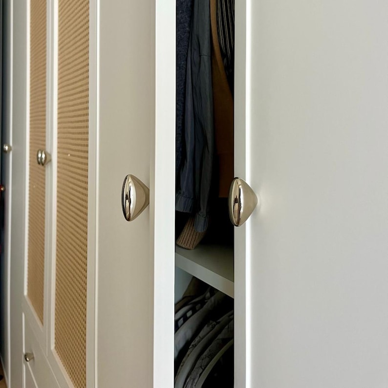 May include: A white wardrobe with gold-colored, cone-shaped knobs. The wardrobe doors are partially open, revealing clothing inside. Some doors have a woven, light brown panel design.