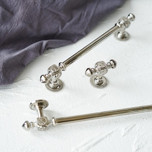 May include: A set of silver-coloured cabinet hardware, including a long handle, a small knob, and a towel bar. The hardware has decorative detailing on the ends and bases, with a classic, vintage design. Suitable for kitchens or bathrooms.