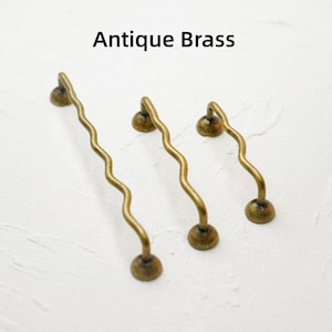 K&ouml;nnte beinhalten: Drei antike Messing-Schrankgriffe mit gewelltem Design. Jeder Griff hat einen runden Sockel und einen gebogenen Griff, mit dem Text "Antique Brass" dar&uuml;ber. Die Griffe sind in verschiedenen Gr&ouml;&szlig;en erh&auml;ltlich.