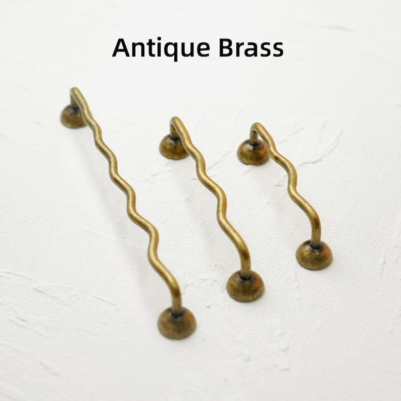 K&ouml;nnte beinhalten: Drei antike Messing-Schrankgriffe mit gewelltem Design. Jeder Griff hat einen runden Sockel und einen gebogenen Griff, mit dem Text "Antique Brass" dar&uuml;ber. Die Griffe sind in verschiedenen Gr&ouml;&szlig;en erh&auml;ltlich.