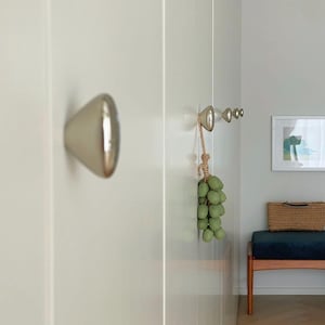 May include: A close-up of a white wall with a silver door knob and a series of silver hooks. A bunch of green decorative grapes hangs from the hooks. A bench with a blue cushion and a woven basket is in the background.