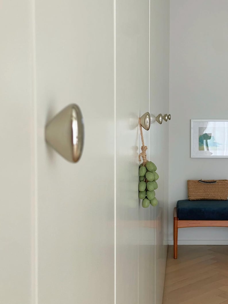 May include: A close-up of a white wall with a silver door knob and a series of silver hooks. A bunch of green decorative grapes hangs from the hooks. A bench with a blue cushion and a woven basket is in the background.