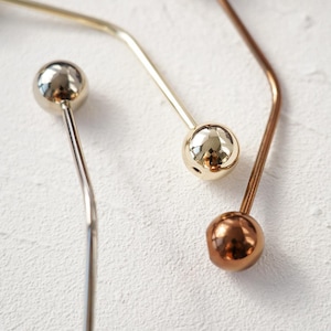 Peut inclure: Gros plan sur trois épingles à cheveux métalliques argentées, dorées et cuivrées. Chaque épingle est ornée d'une tête sphérique et d'une tige incurvée et fine. Les épingles sont disposées en diagonale sur une surface blanche texturée.